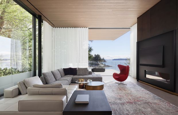 Contemporary waterfront living room with floor-to-ceiling glazing, a large sectional sofa, sculptural red lounge chair, low round coffee table, and a dark built-in fireplace and media wall. The room has a warm timber-lined ceiling, dark window frames, neutral furnishings, and a bright outlook toward the water.