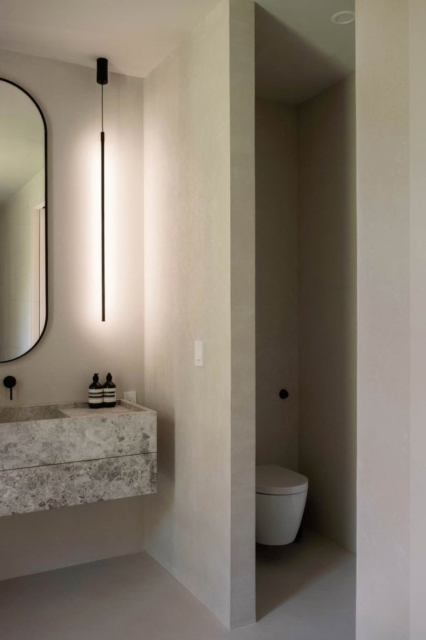 A minimalist bathroom with a floating stone vanity and integrated drawers, a large rounded-rectangle black-framed mirror, a slim vertical pendant light, and a partially concealed toilet alcove. The space uses warm off-white plastered walls and floor surfaces with a pale veined stone vanity, creating a refined, subdued atmosphere.