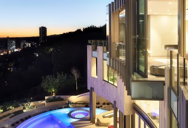 Twilight exterior view of a multi-level contemporary residence on a sloped urban site, with extensive floor-to-ceiling glazing, projecting vertical facade fins, illuminated interiors, landscaped terraces, and a glowing swimming pool below. City lights and a wooded hillside are visible beyond the property.