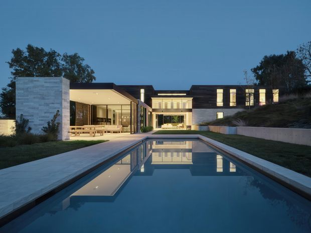 Twilight exterior view of a large contemporary residence arranged around a long rectangular swimming pool. The home features low, horizontal flat-roofed volumes, extensive illuminated glazing, dark roof and trim elements, pale stone-clad walls, and landscaped sloping grounds. A covered outdoor dining or entertaining area opens toward the pool, while the pool provides a strong reflective foreground.