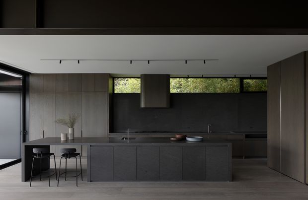 A sophisticated contemporary kitchen with a long central island, integrated dark cabinetry, a matching dark splashback, and a slim linear track light. Floor-to-ceiling glazing and clerestory windows bring in views of greenery, while two black bar stools sit at the island. The palette is restrained and moody, with grey-brown timber cabinetry, charcoal surfaces, and a light grey floor.