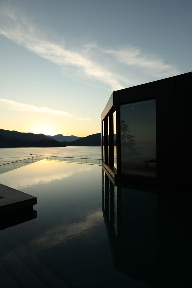 A dramatic dusk exterior photograph of a minimalist, dark-clad modern building beside a calm lake. Floor-to-ceiling glazing reflects the surrounding landscape, while an infinity-edge pool or water terrace extends toward the lake. The scene includes a slim glass balustrade, mountain silhouettes, and warm sunset light creating a serene, high-contrast atmosphere.