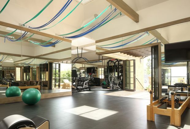A bright, spacious home gym with dark flooring, extensive mirrored walls, exposed timber ceiling beams, large glazed doors and windows, and a variety of strength-training equipment. Green and blue suspension or resistance straps are draped across the ceiling, with exercise balls and wooden-framed equipment visible in the foreground. The room has a high ceiling, abundant daylight, and a contemporary fitness-studio atmosphere.