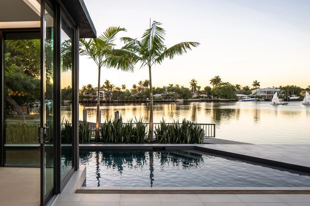 A contemporary waterfront residential setting viewed from a covered indoor-outdoor area, with floor-to-ceiling glazed doors opening onto a narrow swimming pool and landscaped planter. Beyond the pool is a calm canal or lagoon with palm trees, waterfront homes, boats and small sailboats. The scene has clean horizontal lines, dark slim window frames, pale tiled paving and warm sunset light.