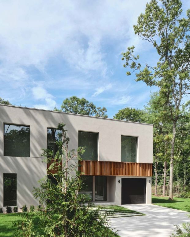Exterior view of a contemporary two-storey house set within a leafy landscaped site. The building has a simple flat roof, smooth light-grey rendered walls, large dark-framed windows, and a projecting upper-level balcony with vertical timber screening. A ground-level garage and paved driveway are visible.