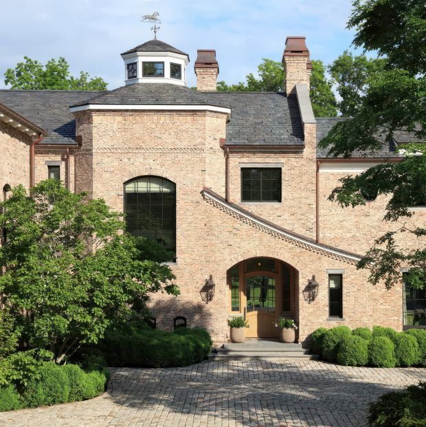 Exterior view of a substantial traditional brick building with a slate roof, prominent arched entry, black-framed windows, multiple chimneys, and a small rooftop cupola topped with a weather vane. Formal clipped hedges and a paved courtyard frame the entrance.