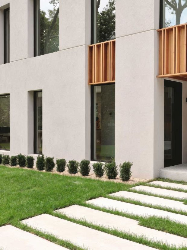 Exterior view of a contemporary multi-storey home with a smooth pale rendered facade, tall dark-framed windows, recessed dark entry door, and warm timber vertical screening panels. A manicured lawn, clipped shrubs, and large rectangular stepping stones form the foreground walkway.