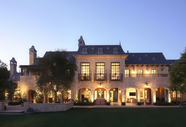 Exterior dusk view of a grand château-style building with symmetrical pale masonry walls, multiple steep dark slate roofs, ornate tower elements, tall illuminated multi-pane windows, arched ground-floor openings, balconies, and landscaped lawns and courtyards in front.