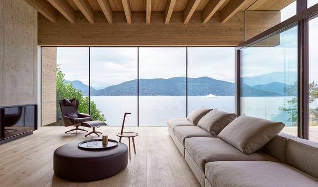 A refined contemporary living room with a large sectional sofa, round ottoman coffee table, lounge chair, and minimalist side table. Warm timber flooring, exposed timber ceiling beams, and timber wall surfaces frame expansive floor-to-ceiling glazing overlooking a calm body of water and mountainous landscape. A dark built-in fireplace or media wall is visible at left.