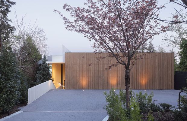 Exterior view of a contemporary single-storey residence, with a large vertically slatted timber-clad facade set behind a broad paved driveway or forecourt. Warm concealed lighting washes the timber wall, while a white angular roof or canopy and entry path are partially visible to the left. Mature garden planting and a deciduous tree frame the building.