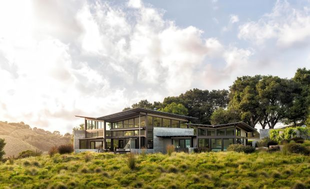Exterior view of a low-profile contemporary home set into a grassy, sloping landscape. The building features extensive floor-to-ceiling glazing, dark horizontal rooflines, timber or dark metal framing, and a combination of solid masonry and glazed volumes. Mature trees and rolling terrain surround the house.