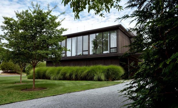 Exterior view of a modern elevated two-storey home set within a landscaped suburban garden. The building has a long rectangular form, dark timber-clad lower level, extensive floor-to-ceiling glazing on the upper level, and a low-pitched flat or near-flat roof. A pale gravel driveway curves through the foreground, with mature trees and ornamental grasses framing the facade.