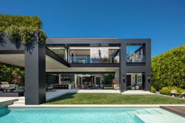 Exterior view of a large contemporary two-storey residence with a dark rectilinear facade, extensive glazing, deep covered terraces, landscaped lawn, and a swimming pool in the foreground. Interior living spaces are visible through the open glazing, and the building has a flat roof with strong cantilevered forms.