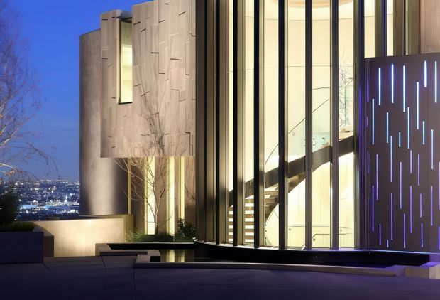 Twilight exterior view of a highly contemporary multi-level residence on a sloping urban site. The building combines tall floor-to-ceiling glazed openings, pale textured solid wall panels, dark vertical framing, and a sculptural illuminated privacy screen with narrow blue light slots. A terrace or roof deck occupies the foreground, while city lights are visible in the distance.