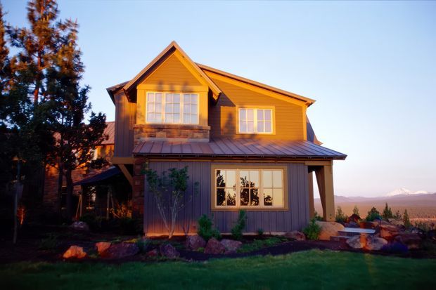 Exterior view of a warm-toned two-storey timber-clad house at sunset. The home has a prominent front-facing gable, pitched rooflines, multi-pane windows, a covered lower-level roof or porch, landscaped planting and rocks around the base, and distant mountains visible beyond the property.