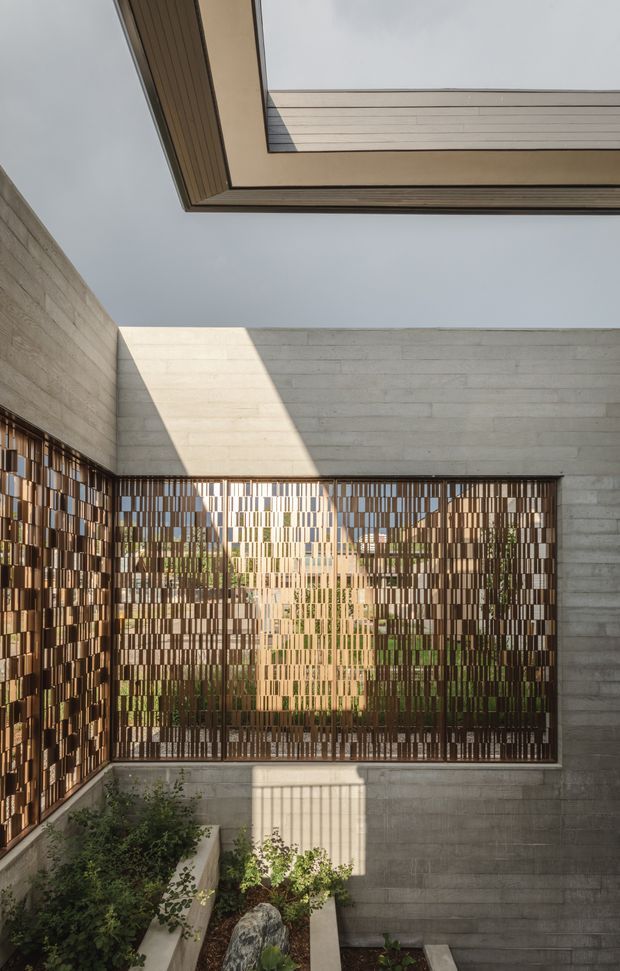 A tranquil contemporary courtyard viewed from inside a concrete-walled space. A large perforated metal screen with a varied vertical and rectangular pattern filters sunlight and views of planted greenery beyond. The courtyard is surrounded by pale horizontal board-formed concrete walls, with raised planter beds, gravel, and a roof opening or skylight above. Strong geometric shadows and warm reflected light create a calm, architectural atmosphere.