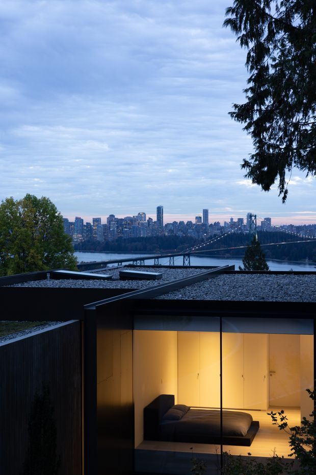 Twilight view of a modern flat-roofed residential structure with a gravel roof and extensive floor-to-ceiling glazing. A warmly lit minimalist bedroom is visible through the glass, while a river, suspension bridge, and dense city skyline form the background. Dark exterior cladding and strong horizontal roof planes create a restrained contemporary composition.