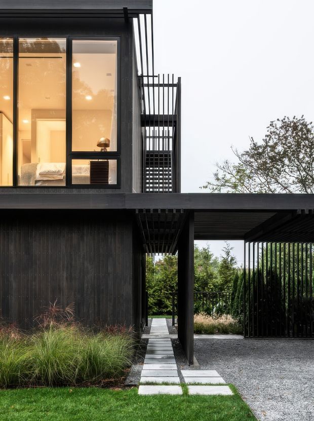 A contemporary dark-clad two-storey house viewed from the landscaped front garden. The facade combines black vertical cladding, large illuminated windows, and strong linear metal screens and balustrades. A covered carport occupies the lower right, while a narrow stepping-stone path leads through ornamental grasses and planting toward the entry. The composition emphasizes crisp geometric lines, open outdoor circulation, and a restrained dark palette against greenery.