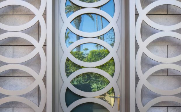 Close-up of a decorative residential entry door or screen assembly with repeated white interlocking oval forms over glazed panels. The central glazing reflects blue sky, palm trees, and lush greenery, while the side panels combine the same geometric white motif with pale textured surfaces. A slim metallic door handle is visible at lower right. The design is highly geometric and ornamental, with strong contrast between white framing, grey panels, and reflective glass.