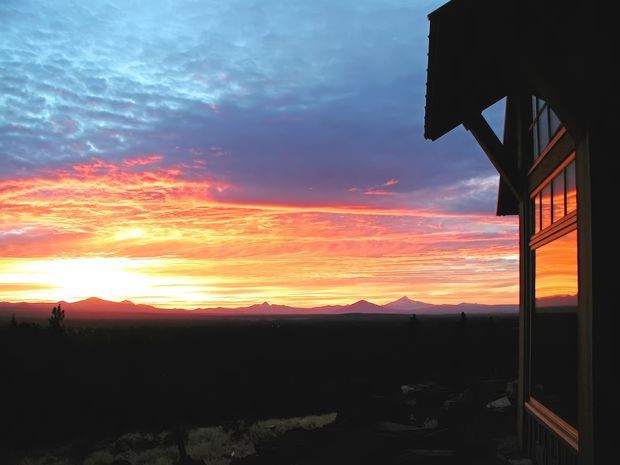 A dramatic sunset landscape viewed beside the dark silhouette of a building. The right side shows an exterior wall with vertical timber-like framing, projecting roof eaves, structural braces, and warm reflections in tall windows. A broad open landscape and distant mountain range stretch across the horizon beneath vivid orange, pink, purple, and blue clouds.
