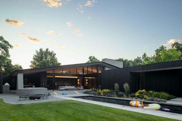 Exterior view of a low-slung contemporary residence with dark vertical cladding, broad floor-to-ceiling glazing, and a shallow pitched roof. The house opens onto a paved entertaining terrace and long narrow reflecting pool, with landscaped garden beds and a manicured lawn in the foreground. Warm interior lighting and outdoor seating are visible through the glazing, creating a refined indoor-outdoor connection.