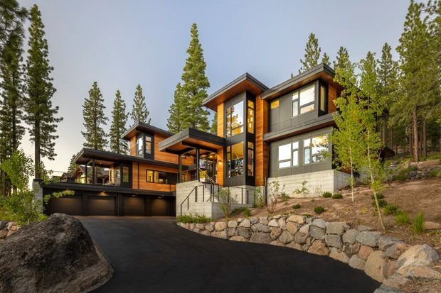 Exterior view of a contemporary multi-level mountain house set into a wooded, sloped site. The home features dark horizontal cladding, warm timber accents, extensive glazing, projecting flat roof forms, a covered entry, and an attached multi-bay garage. A dark asphalt driveway, exposed stone retaining walls, and tall conifer trees surround the building.