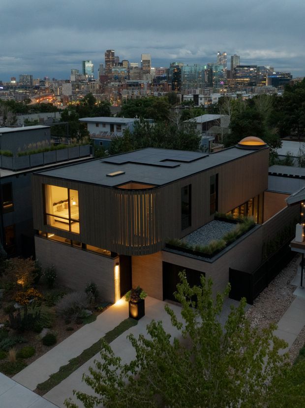 Dusk aerial view of a contemporary multi-level urban residence with a flat roof, dark vertical cladding, warm interior lighting, roof skylights, landscaped roof terraces, and a minimalist concrete-paved entry path. A dense city skyline is visible in the background.