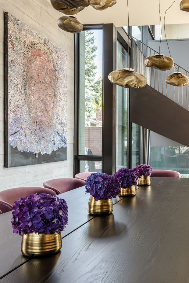 A refined dining area with a long dark timber dining table, rounded upholstered dining chairs, and vivid purple floral arrangements in brass vessels. The room features pale horizontal wall panelling, expansive dark-framed glazing, a sculptural dark stair or balustrade element, and multiple organic-shaped brass pendant lights. An abstract textured artwork adds colour and visual interest.