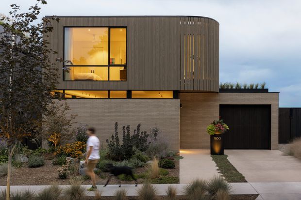 Contemporary two-storey residence viewed from the street at dusk. The upper level has a rounded, vertically clad volume with large black-framed windows glowing warmly from within, while the lower level uses muted masonry and timber-toned cladding. A dark garage door, paved driveway, layered native-style landscaping, and a pedestrian walkway complete the scene.