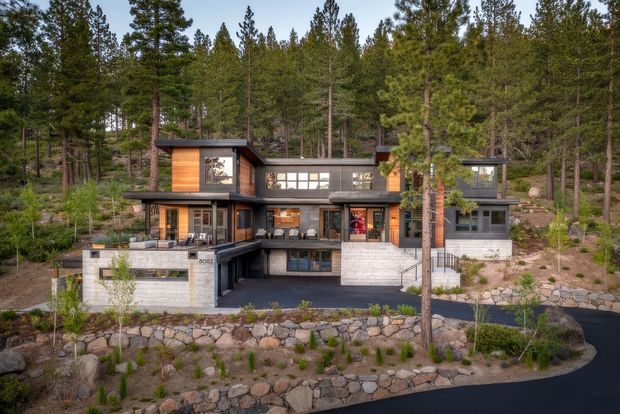 Exterior view of a contemporary multi-level house set into a wooded, sloping site. The building has a dark horizontal facade, warm timber-clad sections, extensive glazing, projecting flat roof planes, balconies and terraces, and substantial stone retaining walls and landscaped paths.