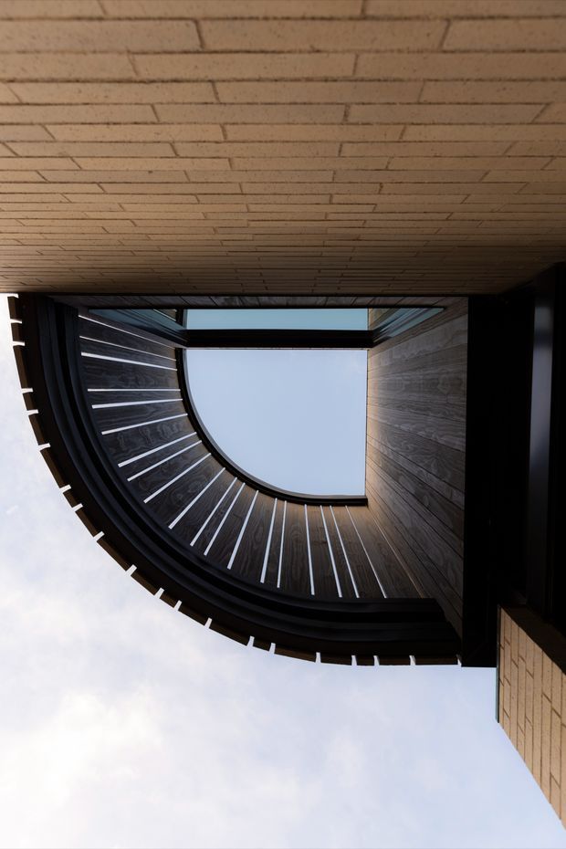 Upward-looking architectural view of a modern building with a dramatic curved, dark timber-clad facade and regularly spaced light-coloured vertical ribs. A pale brick-lined soffit frames the upper edge, while glazing and an open central void reveal the sky. The image emphasizes sculptural geometry, curved rooflines, and contrasting timber, brick, and dark framing.