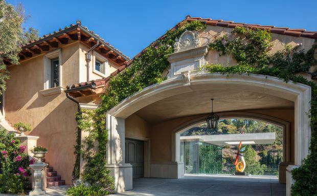 Exterior view of a grand Mediterranean-inspired residence with warm stucco walls, terracotta tiled roofs, ornate white architectural trim, arched porte-cochère, climbing ivy, and landscaped planting. A contemporary glass pavilion is visible through the covered arched passage.