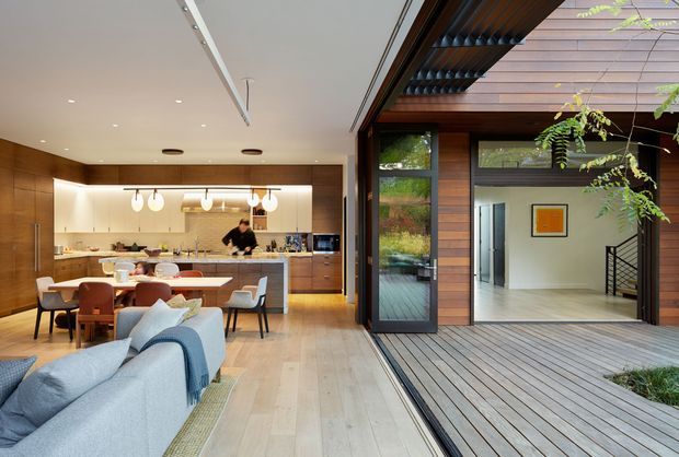 Open-plan contemporary residential interior combining a kitchen, dining area and living room, opening directly onto a timber-lined courtyard deck through large glazed sliding doors. The kitchen features warm wood cabinetry, a pale island benchtop, pendant lighting and integrated appliances. Pale timber flooring continues toward the outdoor deck, while the courtyard side has dark timber wall cladding, overhead timber slats and a visible stair hall beyond.