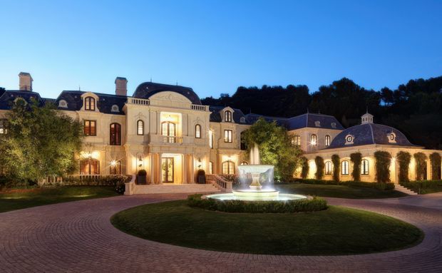 Grand symmetrical mansion exterior photographed at dusk, with an ornate pale stone or stucco facade, multiple wings, steep dark mansard roofs with dormer windows, arched windows and doorways, classical detailing, and a central entrance pavilion. A circular paved driveway and landscaped roundabout with a lit fountain occupy the foreground.