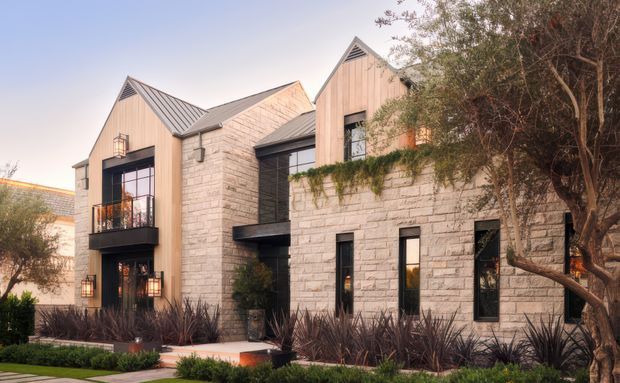 Exterior view of a contemporary multi-storey residence composed of pale masonry walls, warm vertical timber-clad gabled volumes, dark metal-framed glazing and balconies, and pitched standing-seam metal roofs. The building is set within a landscaped garden with mature trees and ornamental planting.