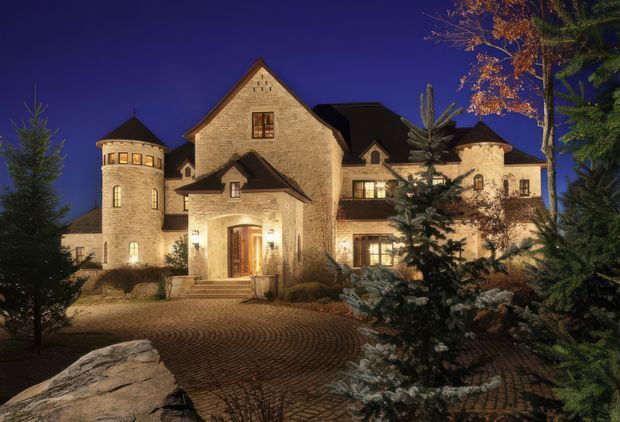 Night-time exterior view of a large stone-clad residence with a steep central gable, multiple pitched roof sections, a round turret, dormer-like projections, and warmly illuminated windows and entry. The building is surrounded by mature evergreen and deciduous landscaping, with a broad curved cobblestone-paved approach in the foreground.