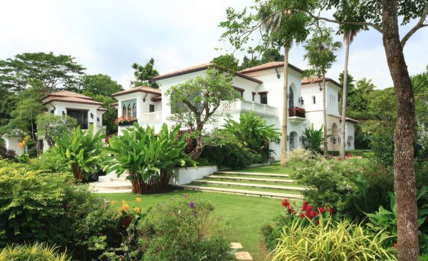 A large white stucco villa set within lush tropical gardens. The building has red clay tiled pitched roofs, arched openings, balconies with decorative balustrades, columns, and multiple connected wings. Broad landscaped steps lead through the garden toward the main entrance, creating a grand resort-like exterior.