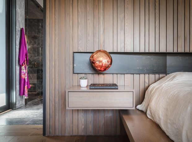 A refined bedroom interior featuring a full-height vertical timber wall and integrated bedside joinery. A low floating drawer, dark horizontal display niche, and suspended copper pendant sit beside the bed, while a bathroom with dark stone finishes is visible through the opening at left. The palette is warm timber, charcoal, copper, and neutral bedding, with a contemporary tailored aesthetic.