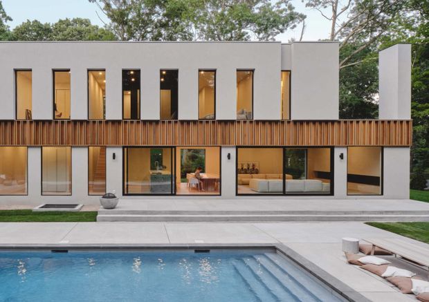 A contemporary two-storey residential facade viewed from the pool terrace. The building has a crisp white rendered exterior, a strong horizontal timber slat balcony or screen across the upper level, tall narrow windows, and extensive ground-floor glazing revealing warm-toned interiors. A swimming pool, paved terrace, loungers, and landscaped garden surround the house.