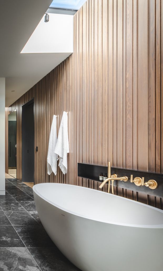 A refined contemporary bathroom featuring a freestanding white oval bathtub, wall-mounted brushed brass tapware, a black horizontal splashback panel, dark stone-look floor tiles, and a full-height vertical timber slat feature wall. A skylight brings natural light into the elongated space, with a dark doorway and towel hooks along the corridor-like side wall.