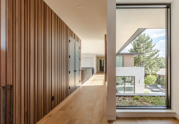 A bright contemporary residential hallway with a long wall of vertical dark timber slats and integrated flush doors on the left. The corridor has pale timber flooring, white walls and ceiling, and a large black-framed window overlooking a modern landscaped courtyard and adjacent architecture. A built-in dark kitchen or cabinetry zone is visible farther down the hall.