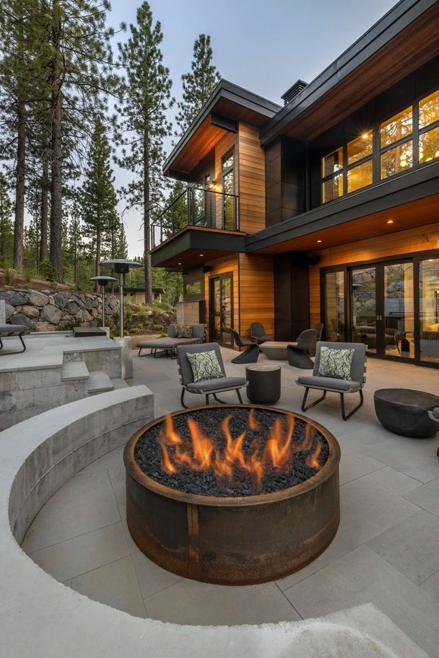 A modern multi-level timber and dark-clad house set among tall pine trees, viewed from a landscaped outdoor terrace. The terrace features pale large-format paving, curved concrete retaining walls and steps, a circular metal fire pit with flames, contemporary lounge chairs, and low outdoor tables. Large glazed doors and windows connect the terrace to the warmly lit interior, while a balcony projects above.
