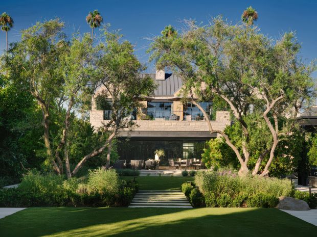 Exterior view of a multi-storey contemporary residence set within a lush landscaped garden. The home features pale masonry walls, dark metal-framed glazing, a steep central metal roof element, and a recessed ground-level outdoor living area. A broad manicured lawn, clipped hedges, planted borders, and a formal stepped path lead toward the house, with mature trees and palms surrounding the property.