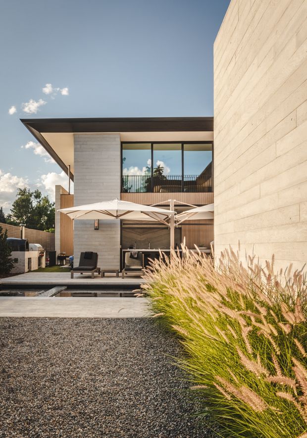 A contemporary two-storey residence viewed from its landscaped outdoor area. The home features broad concrete block walls, timber-clad sections, expansive dark-framed glazing, and a thin projecting flat roof. A paved entertaining terrace with large white umbrellas, outdoor lounge furniture, built-in barbecue, and a narrow reflecting pool sits in front of the house. Ornamental grasses and dark gravel define the foreground garden.