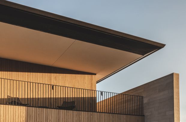Close-up exterior architectural view of a contemporary upper-level balcony beneath a broad projecting roof canopy. The facade is clad in warm vertical timber slats, with slim black metal balustrades and dark roof edging. A smooth grey concrete wall forms the side elevation, creating a minimalist composition against the sky.