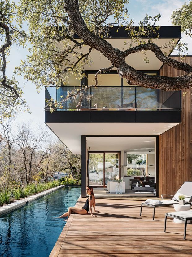 A modern multi-storey residence set among mature trees, viewed from a poolside timber deck. The house features strong horizontal cantilevered volumes, black trim, extensive glazing, a glass-balustraded upper balcony, and vertical timber wall cladding. A long narrow swimming pool runs beside the deck, with an open-plan living area visible through the ground-floor glazing.