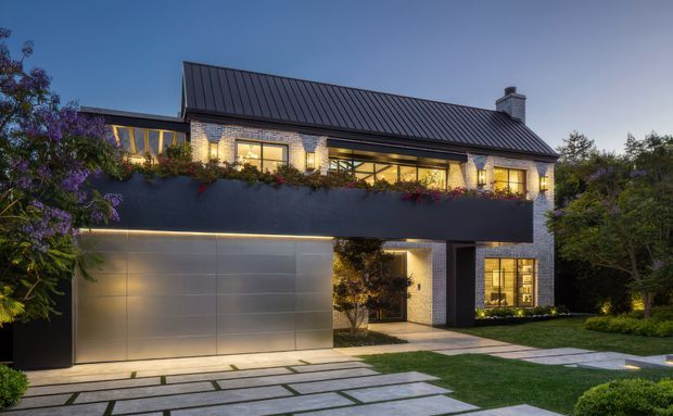 Contemporary multi-storey residence photographed at dusk, featuring a dark standing-seam pitched roof, pale brick masonry, extensive warm-lit glazing, a projecting dark upper volume with planted balcony edge, and an integrated double garage. Large-format pale paving leads through a landscaped front garden to the recessed entry.