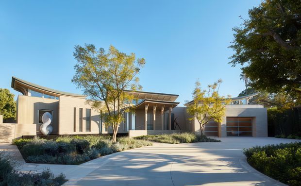 Contemporary single-storey residential house viewed from the driveway, featuring a low horizontal composition, light rendered walls, broad projecting roof eaves, deep covered entry, vertical glazing, and an attached double garage. The front garden uses restrained drought-tolerant planting and paved concrete surfaces.