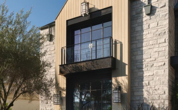 Exterior view of a multi-storey building featuring pale tan vertical cladding, light rough-textured masonry walls, black-framed gridded glazing, and a projecting upper-level balcony with a clear glass balustrade. Black metal lantern-style wall lights flank the entrance and upper opening. Trees partially frame the facade.