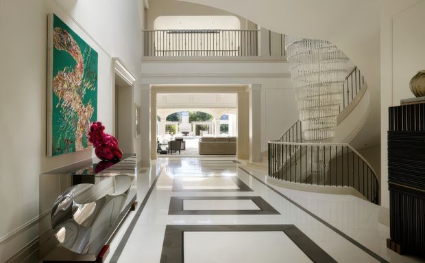 A grand, bright residential entry hall with a double-height void and mezzanine balustrade. The polished light floor features bold dark rectangular inlays, while a sculptural curved staircase with a dramatic tiered pendant chandelier dominates the right side. A console and artwork line the left wall, and a formal living area is visible beyond the hallway.
